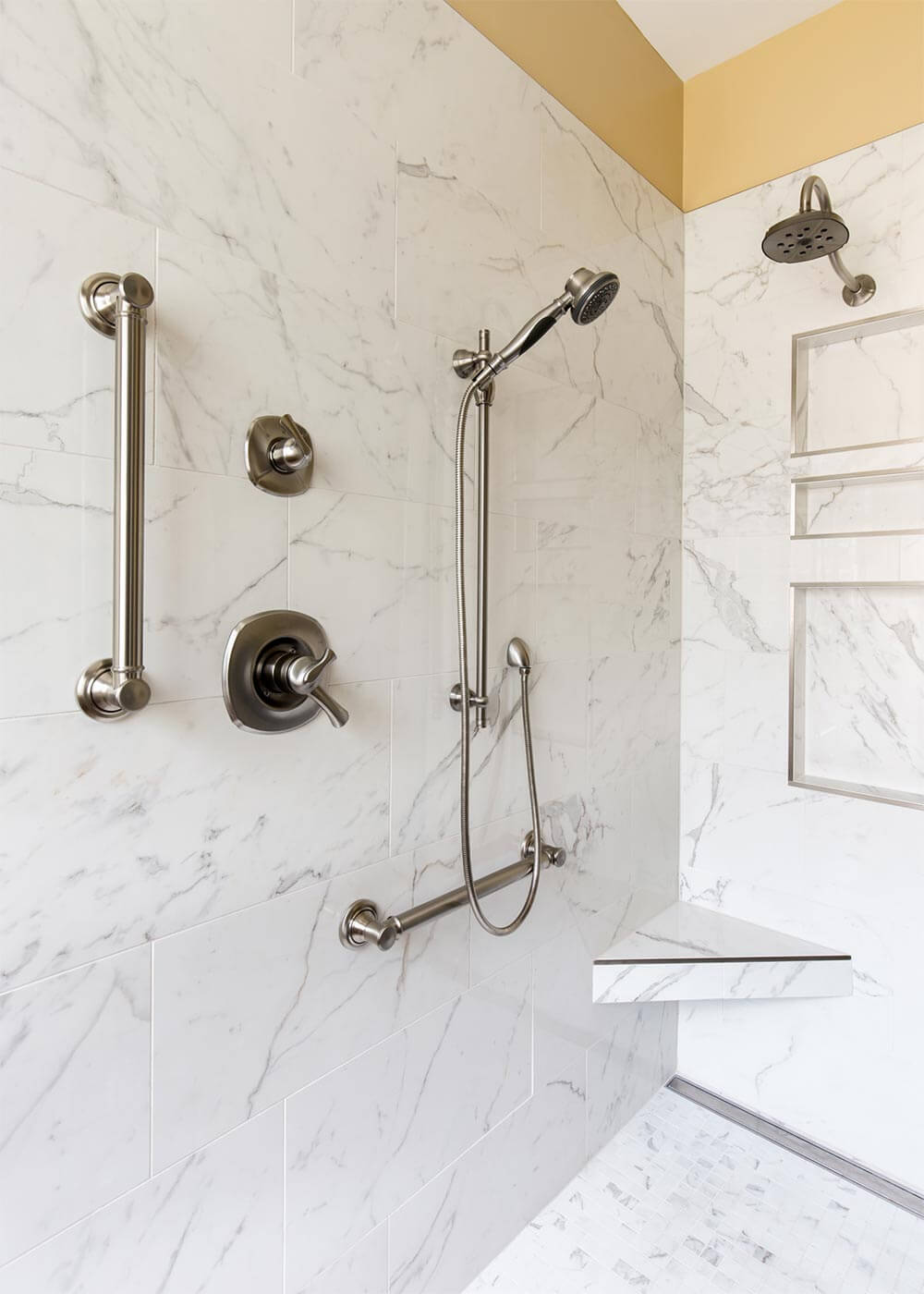 Modern shower with marble tiles, stainless steel fixtures, handheld and overhead showerheads, and safety grab bars. 