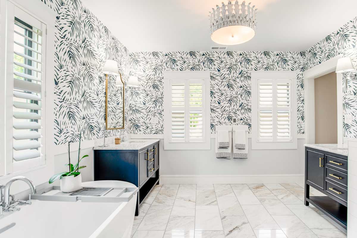 Tropical themed bathroom with palm leaf wallpaper, marble floor, and navy blue vanity units with gold accents. 