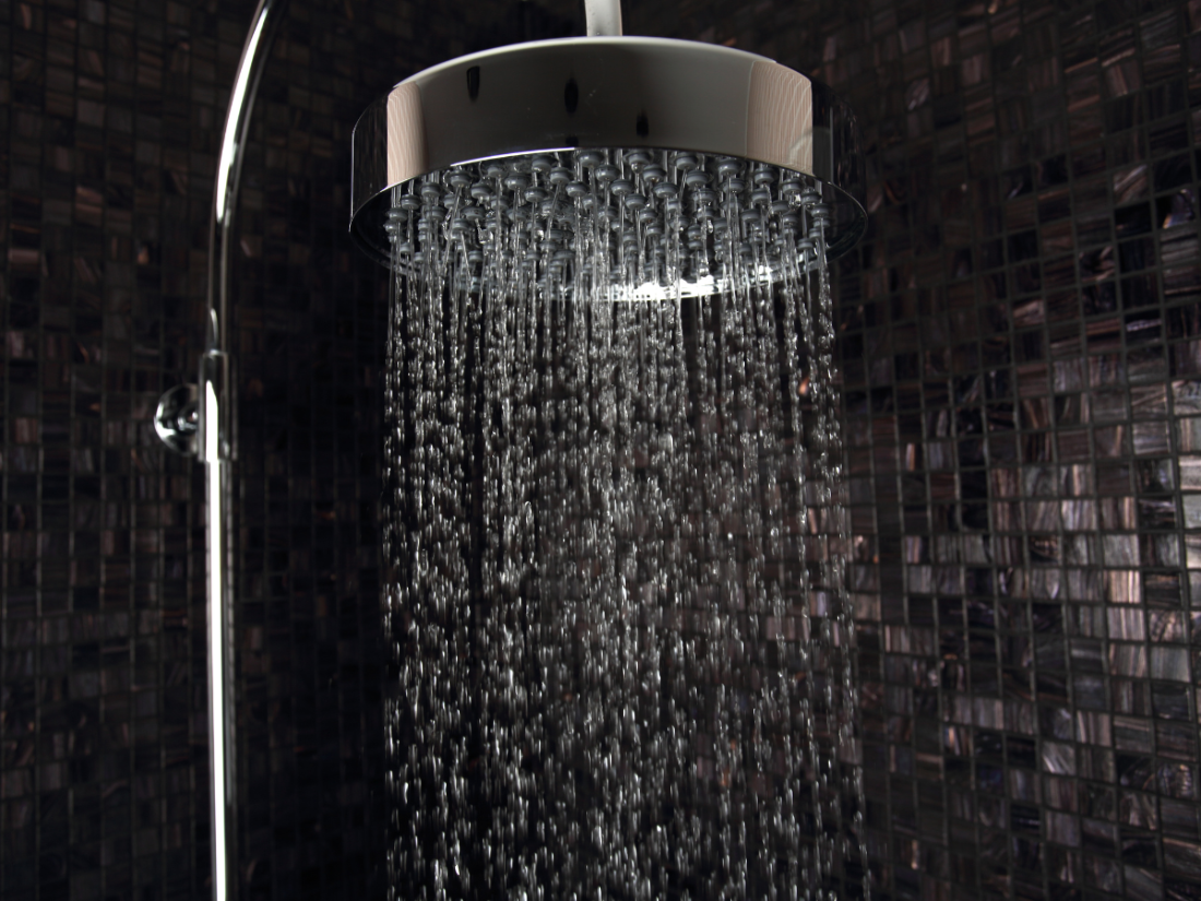 Close-up of a modern rain showerhead with water flowing, set against a dark mosaic tiled wall. 