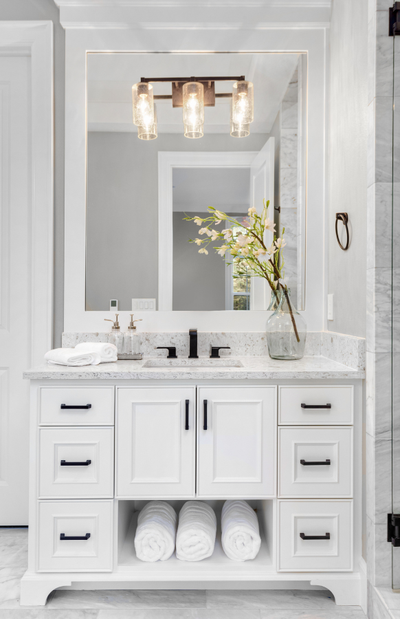 Elegant bathroom vanity with white cabinetry, marble countertop, and illuminated mirror above a vase with fresh flowers. 