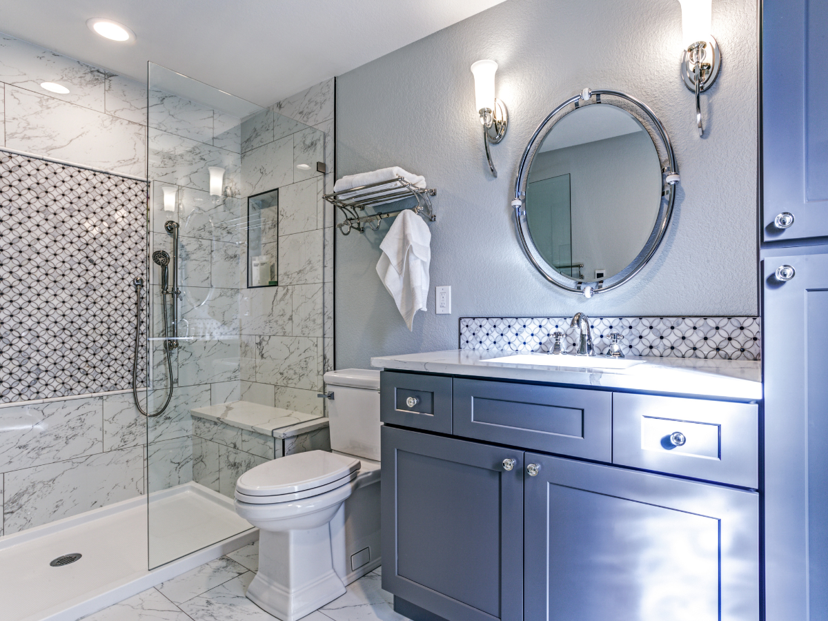 Modern bathroom featuring a navy blue vanity, marble wall tiles, glass shower, and circular mirror with wall sconces. 