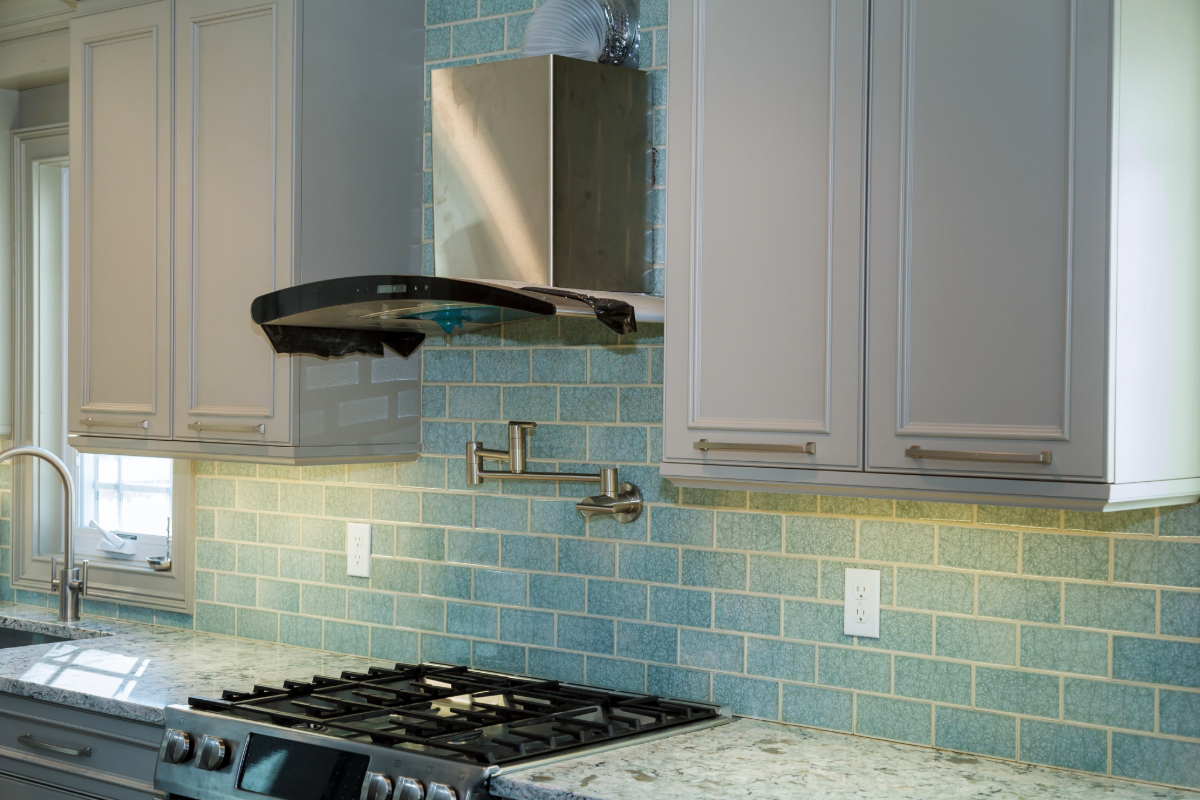 Contemporary kitchen with gray cabinetry and blue tiled backsplash, featuring a stainless steel gas stove and range hood. 