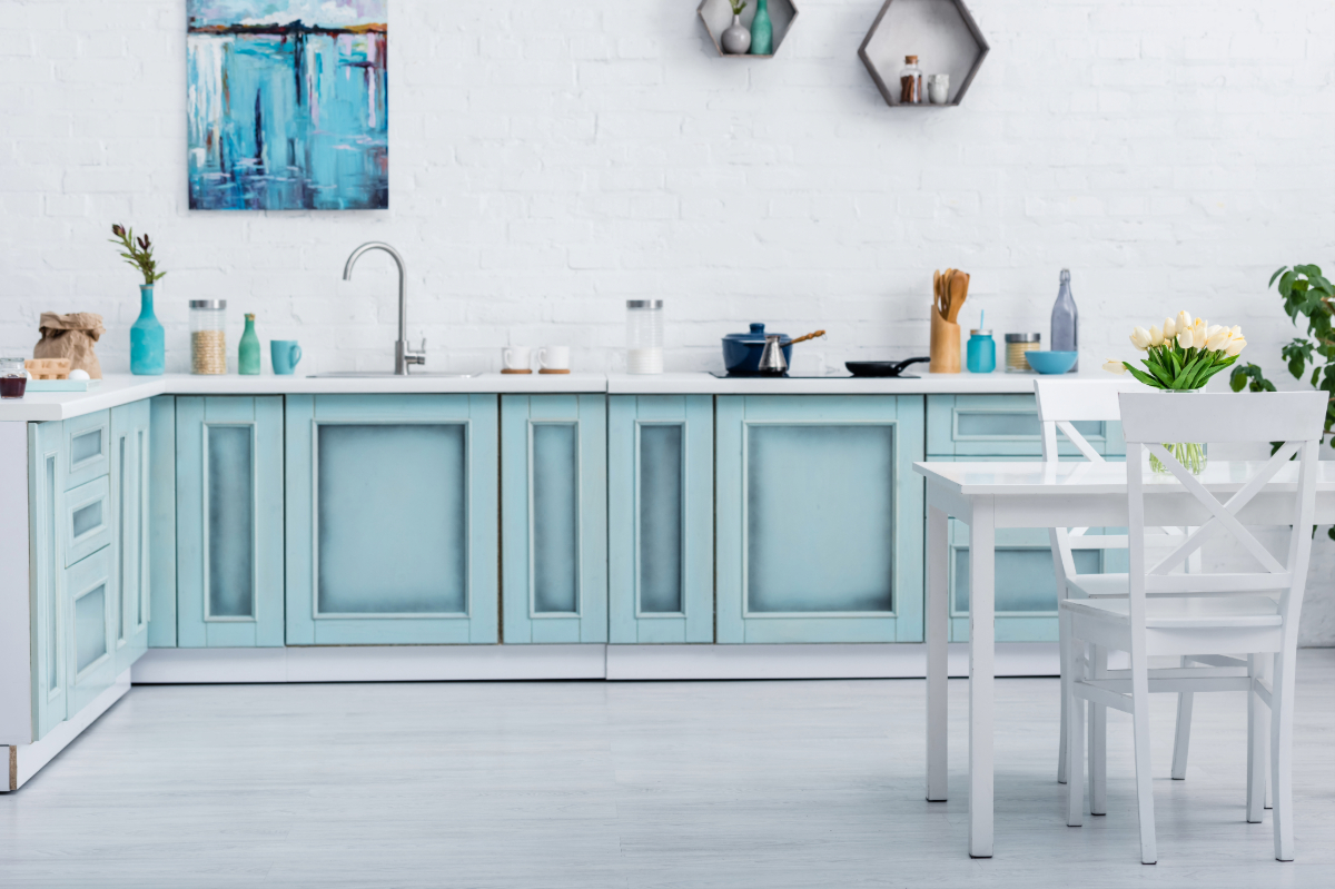 Bright kitchen with light blue cabinetry, white countertops, and dining area, set against a white brick wall with a modern art painting. 