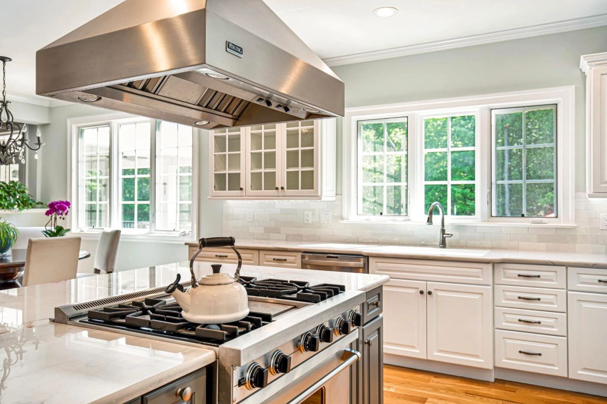 Luxurious kitchen with white cabinetry and marble countertops, featuring a professional gas stove and large stainless steel hood. 