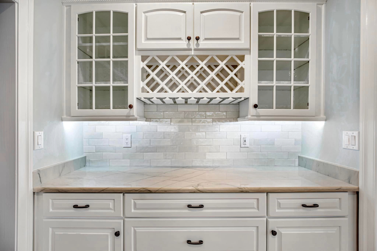 White kitchen nook featuring a built-in wine rack, glass cabinet doors, and a light marble countertop with a tiled backsplash. 