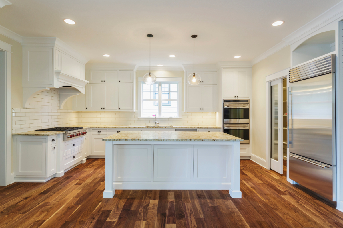 Elegant white kitchen featuring a subway tile backsplash, central island with granite top, and high-end stainless steel appliances & hardwood flooring. 