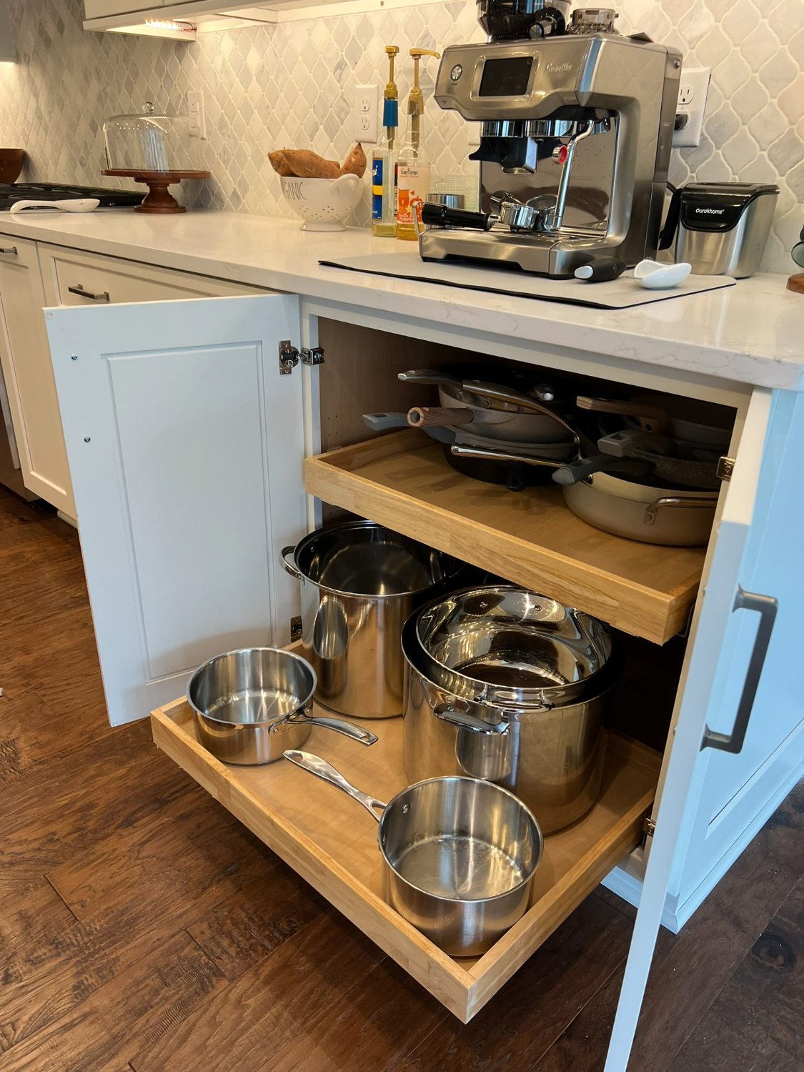 White kitchen cabinet with pull-out drawers showcasing organized pots and pans, next to a countertop with a modern espresso machine. 