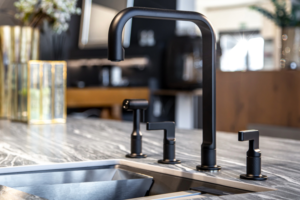 Close-up of a modern matte black kitchen faucet on a marble countertop with a stainless steel sink and decorative glassware. 