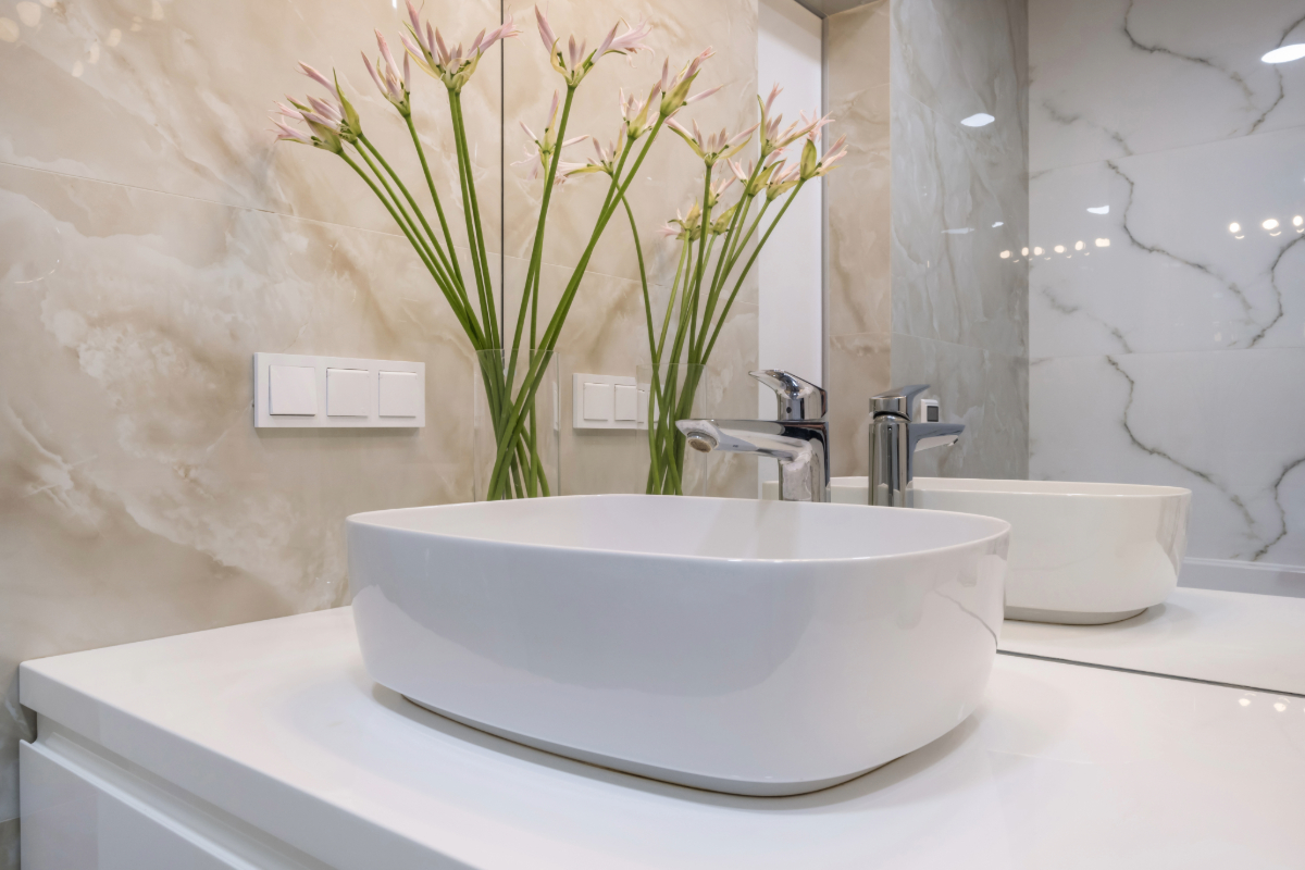 Contemporary bathroom with twin oval basins, marble walls, and a tall vase of pink flowers. 